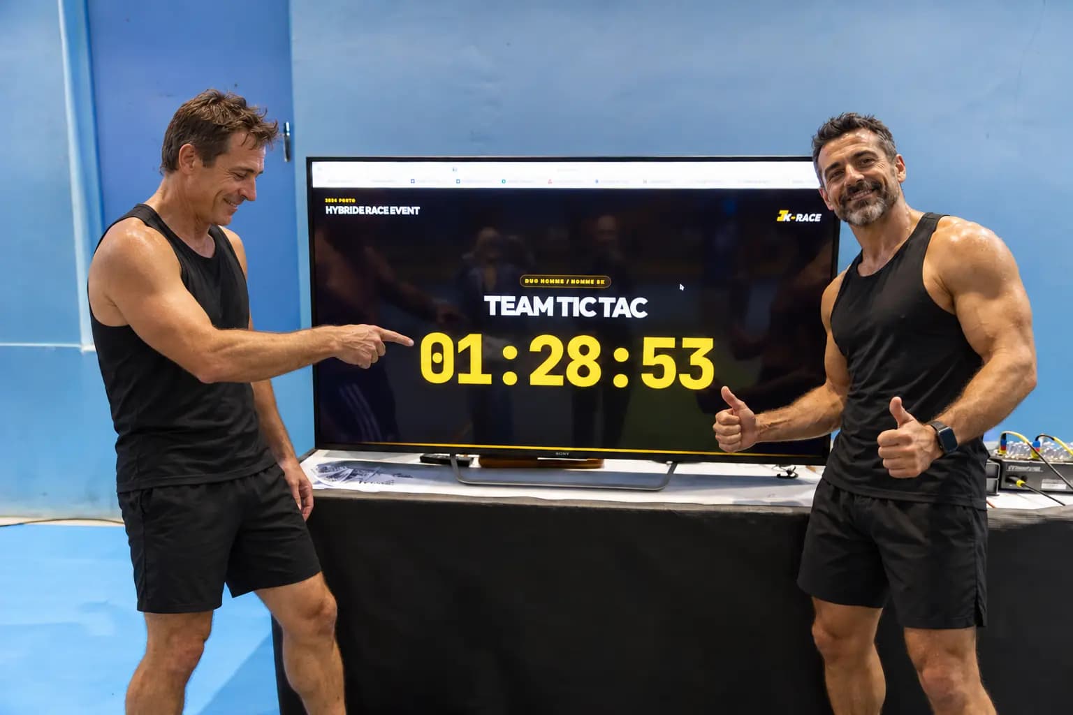 Two athletes of Team Tic Tac pointing at their 01:28:53 time on the Tk-RACE finish board